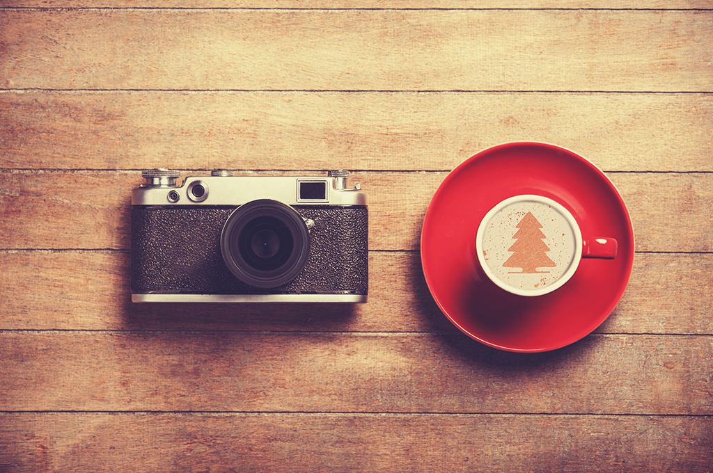 an image looking down on a black and silver vintage camera sitting on a wooden table next to a red cup and saucer of coffee with a christmas tree design on top