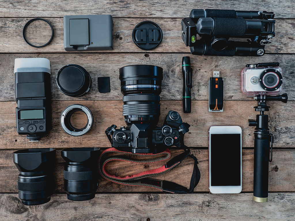 overhead view of photography equipment including digital camera, battery charger, memory card, tripod, flash, action camera, lens and camera accessory on wooden background