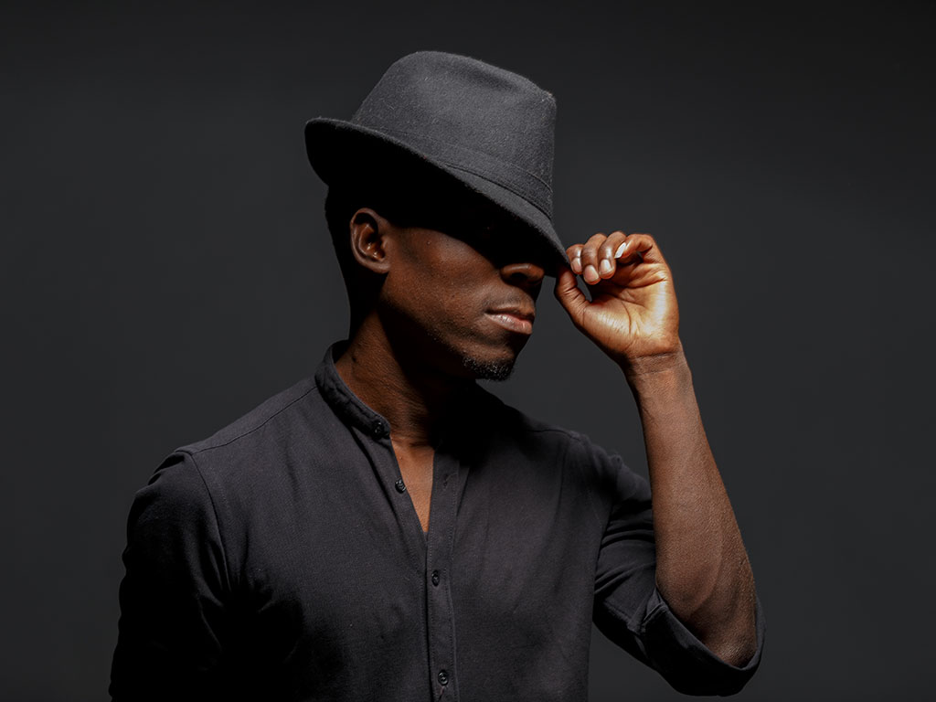 portrait photo of a young black man in a black hat and black shirt against a black background