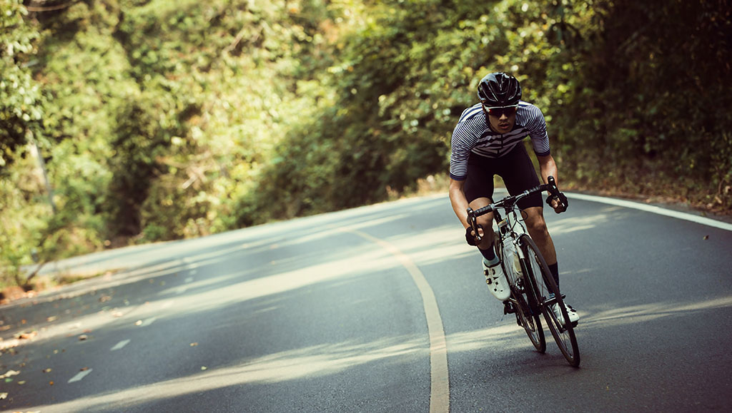 a man in full cycling gear is cycling towards the camera on a road surrounded by trees