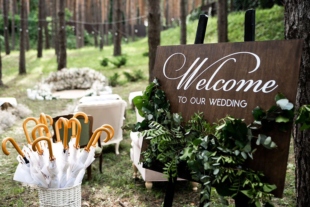 Outdoor wooden welcome board decorated with green branches, basket with umbrellas in case of rain