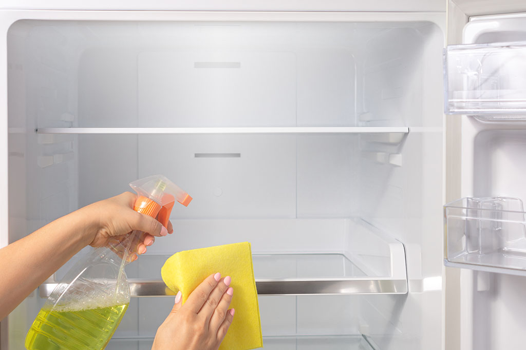 A persons hands holding a spray bottle and cloth cleaning the fridge 