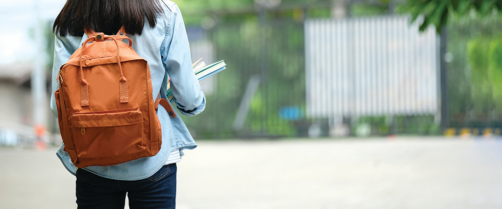 a student standing outside some gates with a rucksack and holding some books 