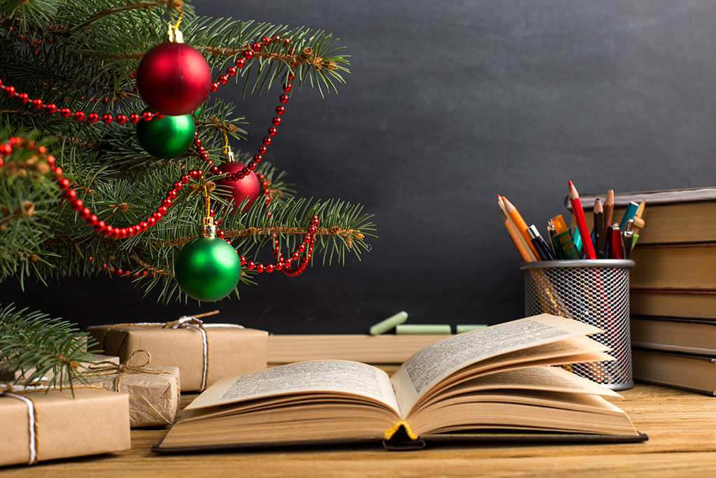 a desk with an open book and a pot of pens, and a Christmas tree with some presents underneath it 