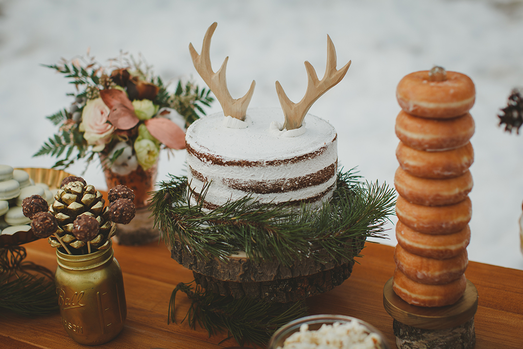 winter wedding idea,  a cake with reindeer antlers on a table with flowers, donuts on a wheel and a golden acron with chocolates on sticks 