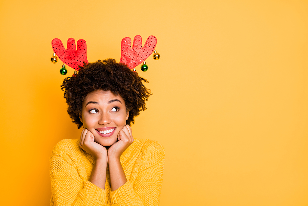 woman with reindeer antlers on her head, smiling and looking to the side with a yellow background that matches her yellow jumper   