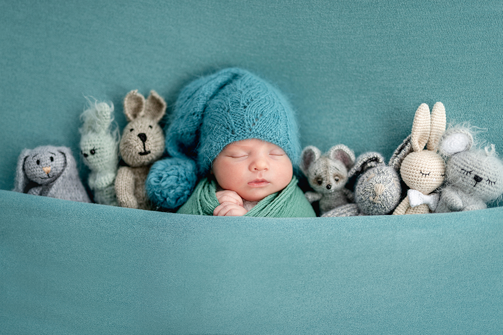A new born baby under a blanket, with a hat, surrounded by toys as a baby photoshoot idea 