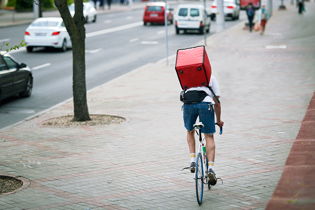 Man delivering food with red thermal backpack, riding bicycle in the city. Food delivery service. Courier on bicycle riding fast through the pedestrian street, deliver food to costumer.