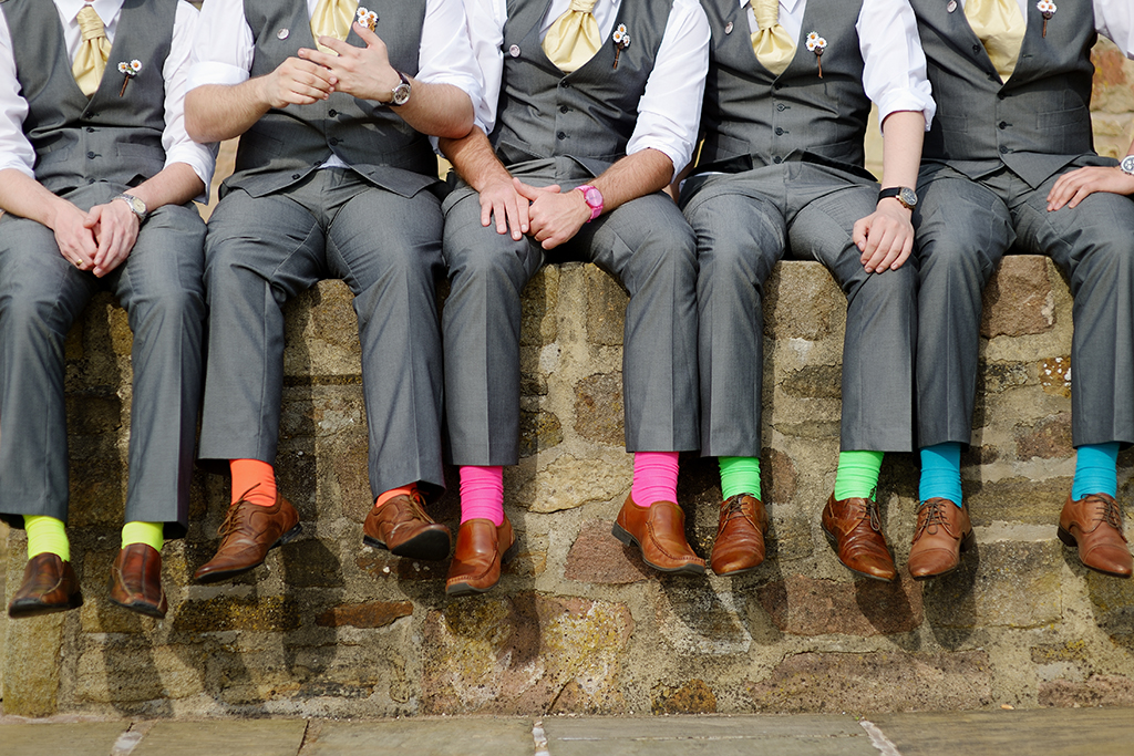 a wedding photography picture idea, a group of groomsmen sitting on a wall showing their brightly coloured shoes in grey suits.